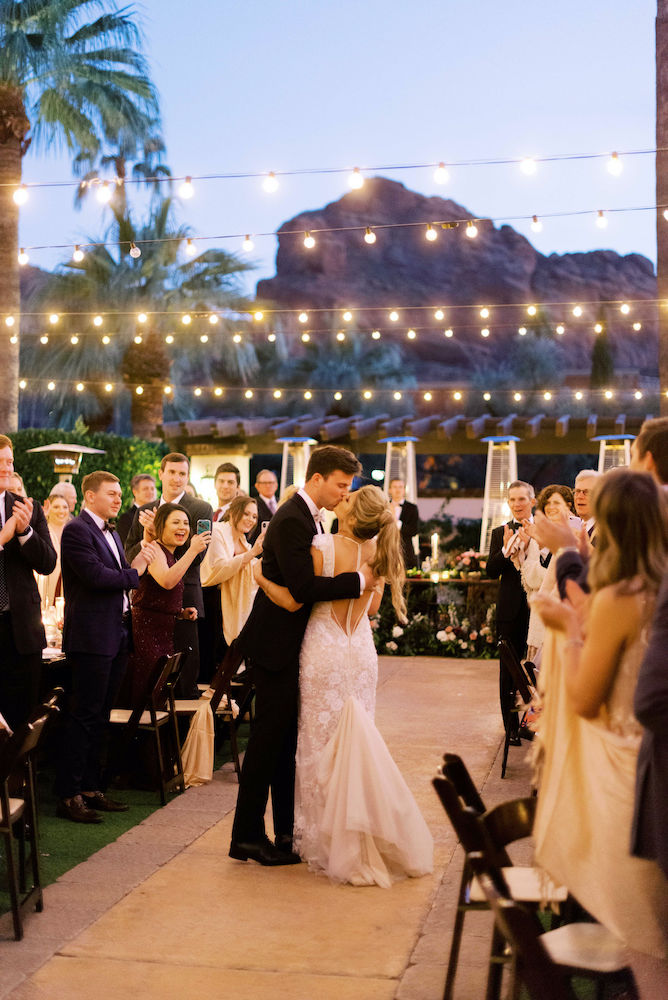 Bride and groom kissing at grand entrance of outdoor wedding reception with bistro lights.